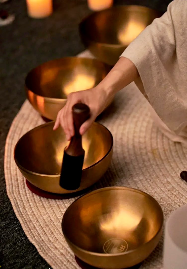 A felt mallet resting in a brass singing bowl in a dim room, a hand still on it, with more bowls waiting beside it