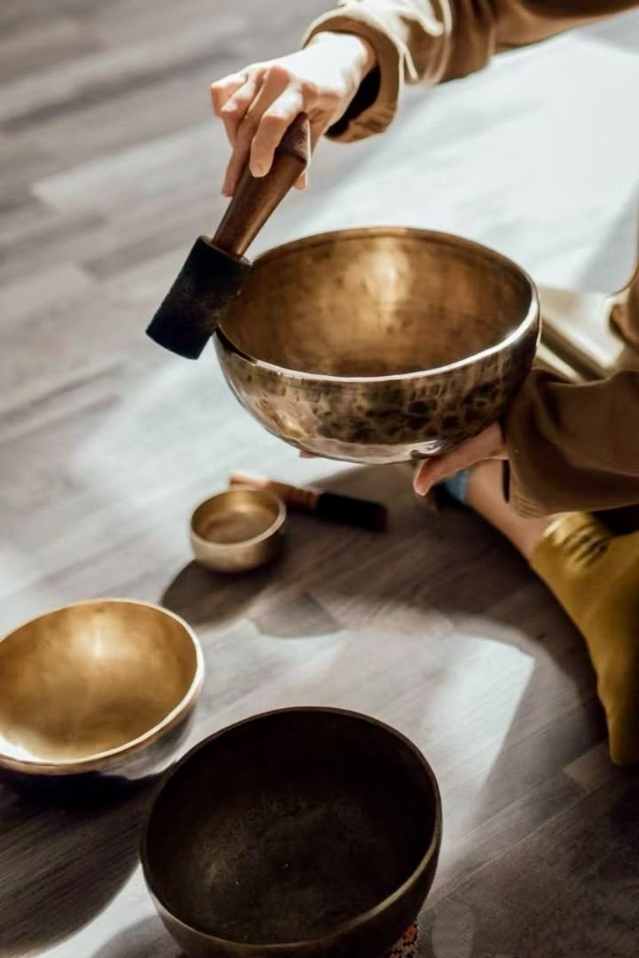 Hands holding a hammered brass singing bowl and bringing a felt mallet to its rim, with three more bowls resting on the floor